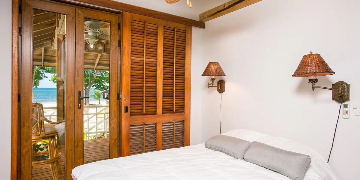 A cozy hotel room with a bed, two wall lamps, and a glass door opening to a balcony with a beach and ocean view.