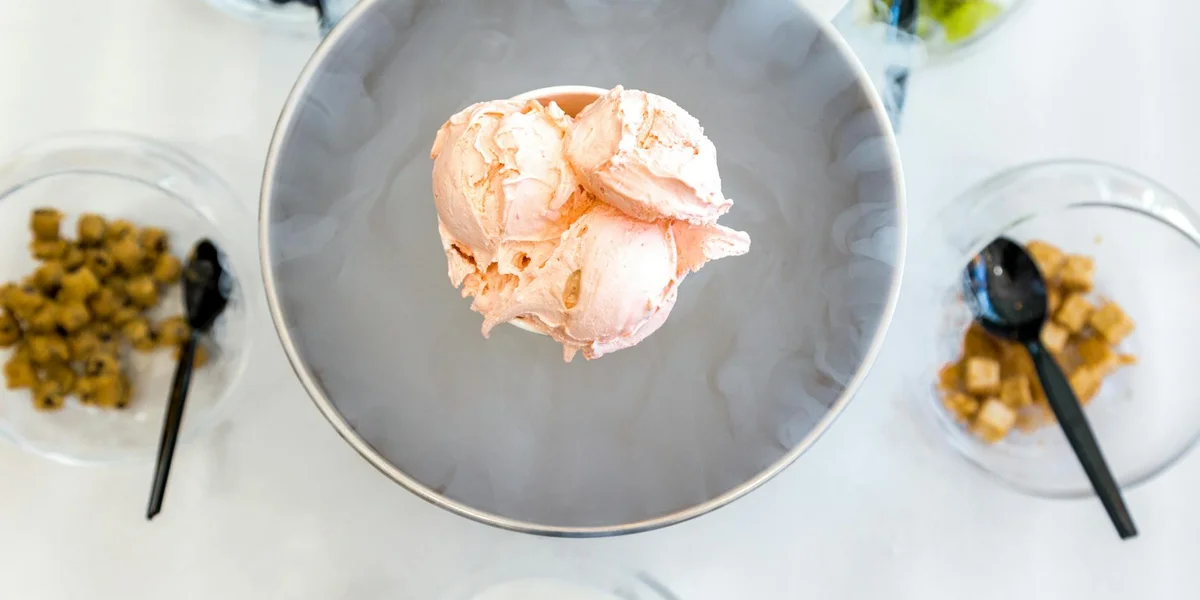 Top-down view of a single scoop of pale pink ice cream centered on a gray bowl, with two side bowls containing crumbled toppings and black spoons nearby.