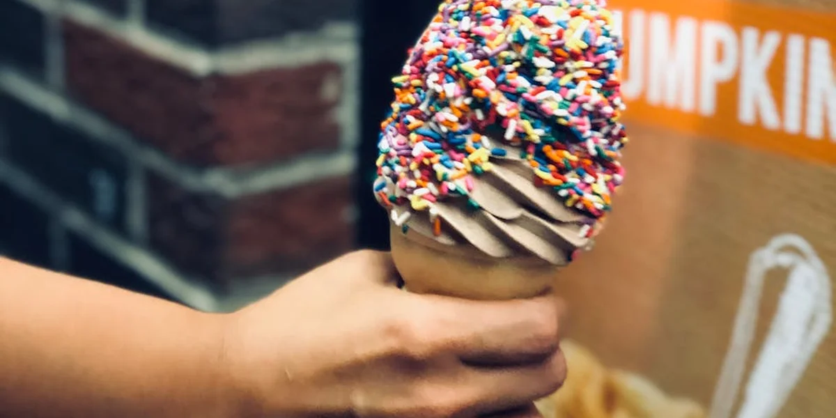 A hand holding a vanilla ice cream cone with rainbow sprinkles against a brick wall