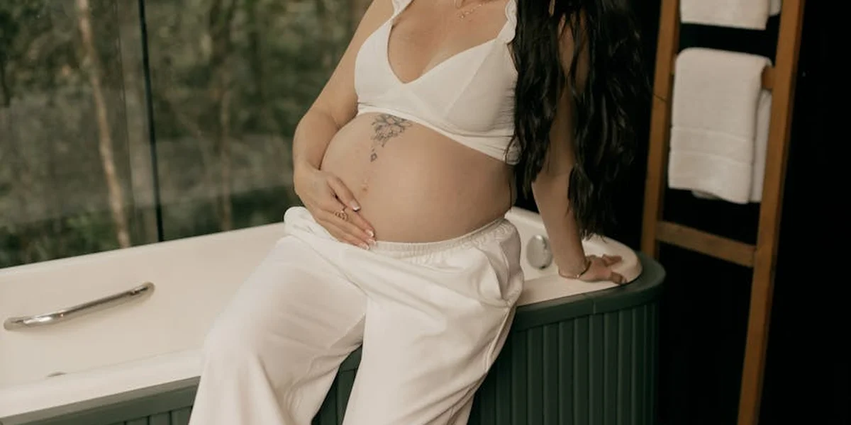 Pregnant woman in white clothing sits by a bathtub, resting her hands on her rounded belly.