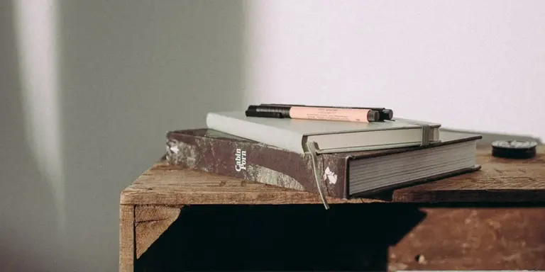 A stack of old books on a rustic wooden desk with a pencil resting on top, suggesting study and reflection.