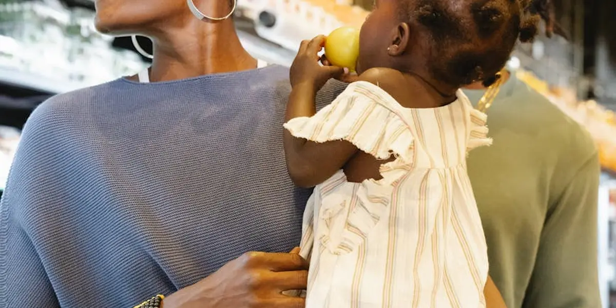 A woman holds a toddler in a grocery store, the child reaching for a fruit as they explore an aisle.