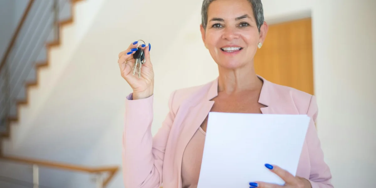 Smiling woman in a light pink blazer holds a key in one hand and a document in the other, standing indoors near a staircase.