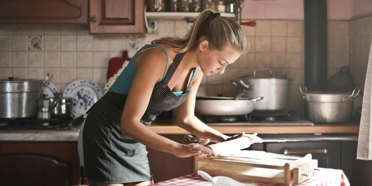 A woman in a kitchen wearing an apron chops food on a wooden cutting board.