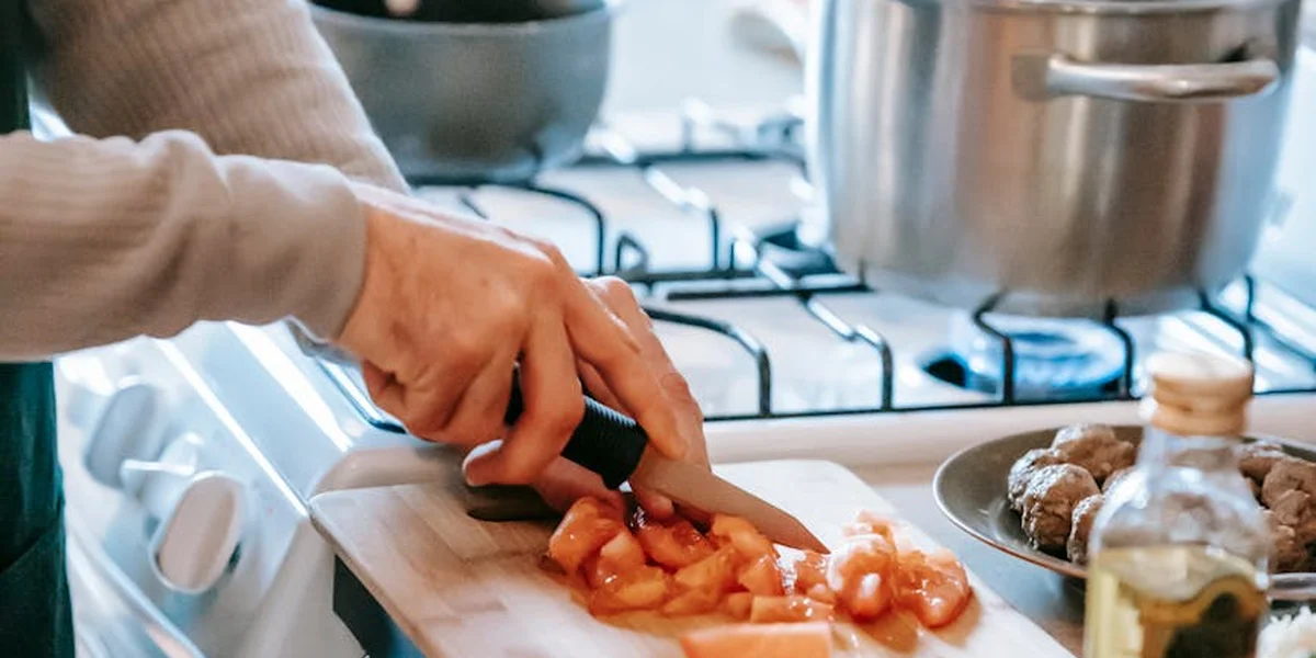 A pair of hands slicing tomatoes on a cutting board in a bright kitchen, with a stove and pot in the background.