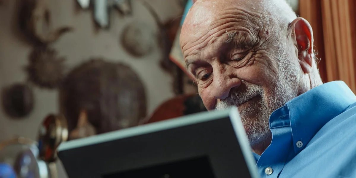 Elderly man in a blue shirt looks at a tablet with a warm, thoughtful smile in a cozy home study.