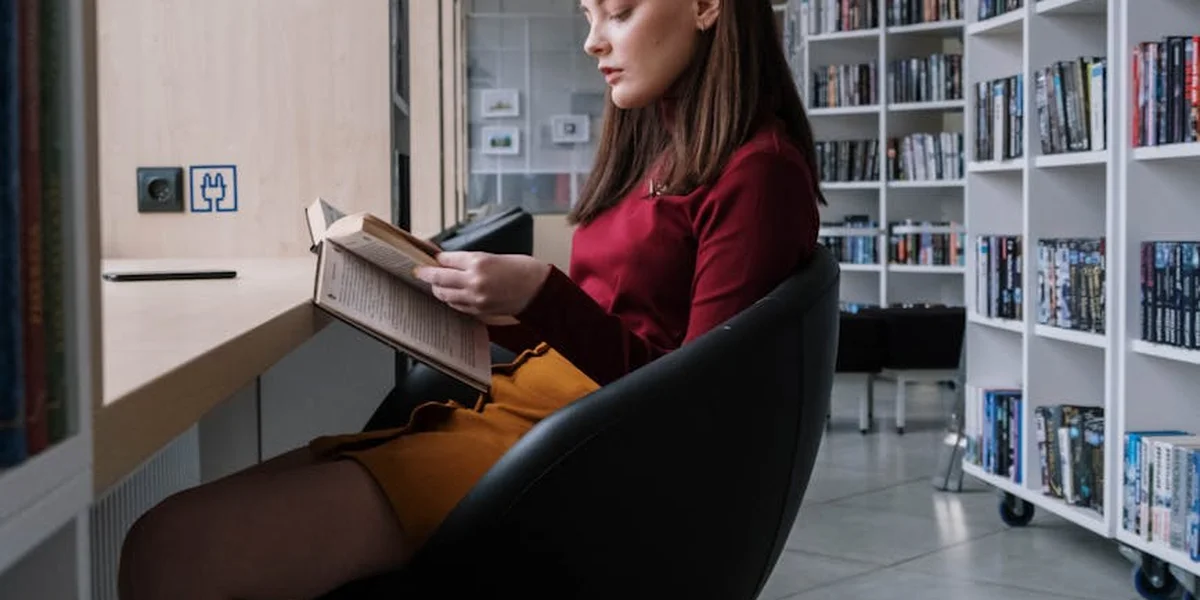 A woman sits in a modern library, reading a book, with tall shelves filled with books around her.