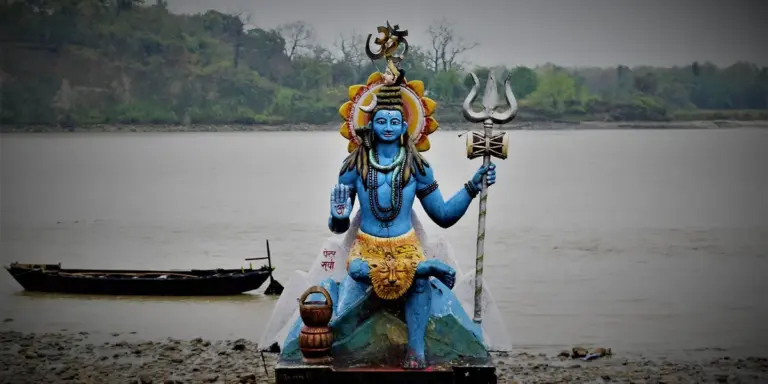Blue-skinned statue of Lord Shiva seated by a river, holding a trident (trishula) and wearing traditional ornaments.