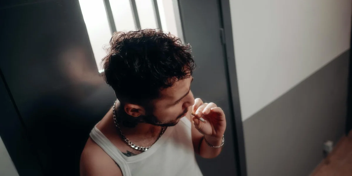 A man in a white sleeveless shirt stands in a dim hallway near an elevator, touching his lips with his fingers.