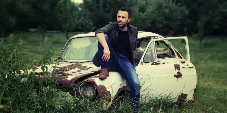 Man wearing casual clothes sits on a rusted vintage car in a green field, with the car's door open and trees in the background.