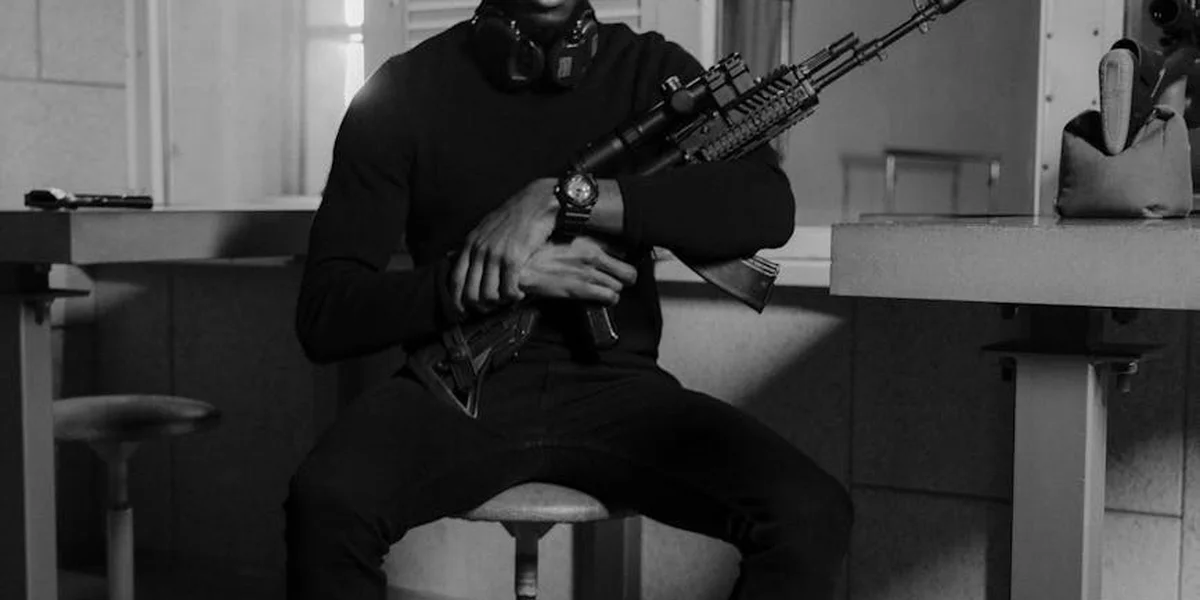 A person wearing a dark hoodie sits on a stool indoors, cradling a rifle with both hands; the image is in black and white.