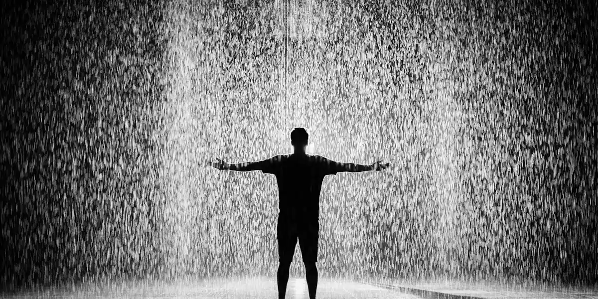 Silhouette of a person standing with arms outstretched beneath a heavy veil of cascading water, evoking immersion in emotional and physical change.