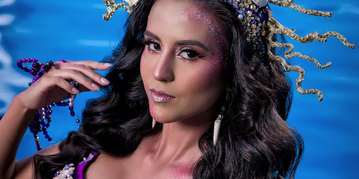 Mermaid-inspired portrait: a woman with shimmering makeup and sea-themed accessories against a blue ocean backdrop.