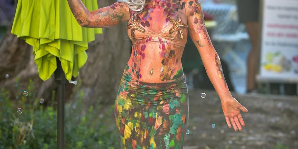 A person wearing vibrant mermaid-inspired body paint and a scale-pattern wrap stands outdoors beside a lime-green umbrella.