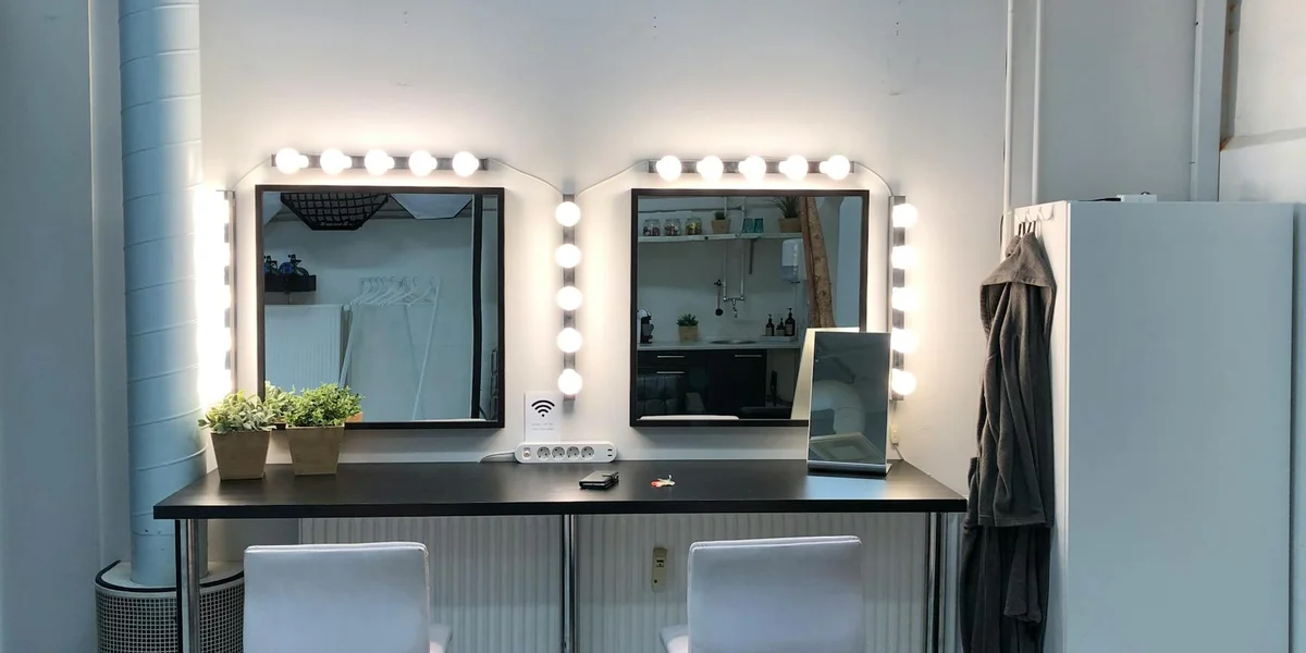 A backstage makeup station with two illuminated vanity mirrors, a plant on the counter, and a reflection of the space, symbolizing transformation and self-reflection.