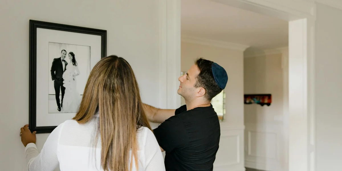 A man wearing a dark shirt and a cap looks up as he adjusts a framed black-and-white wedding photo on a wall, while a woman with long blonde hair stands nearby with her back to the camera in a bright, modern home.