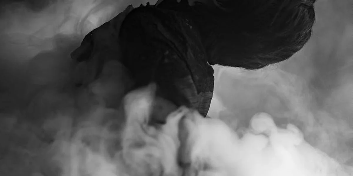 Monochrome photograph of a person bent forward with their head down, enveloped in hazy smoke or fog.