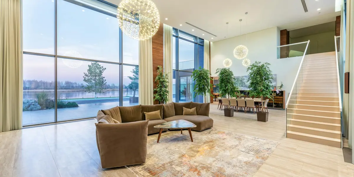 Modern lobby interior featuring a staircase on the right, expansive floor-to-ceiling windows, brown sectional seating, round pendant lights, and potted plants.