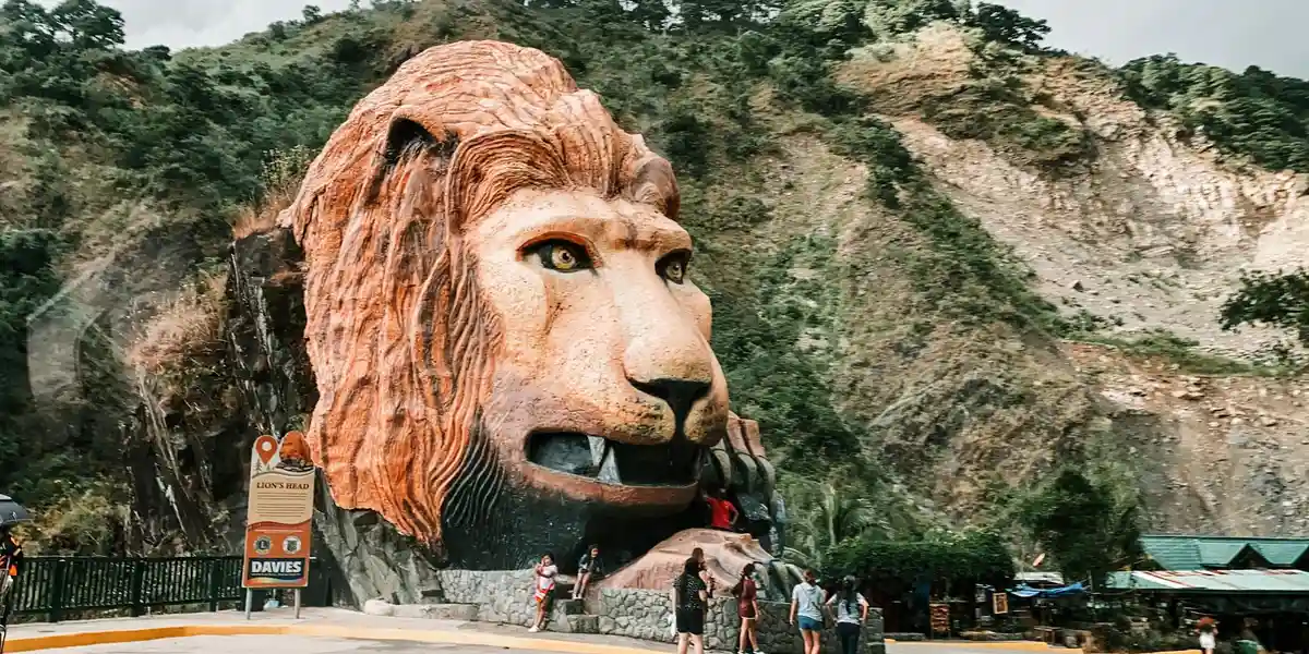 Carved stone mountain lion statue embedded in a rocky hillside, with a few visitors nearby and trees in the background.