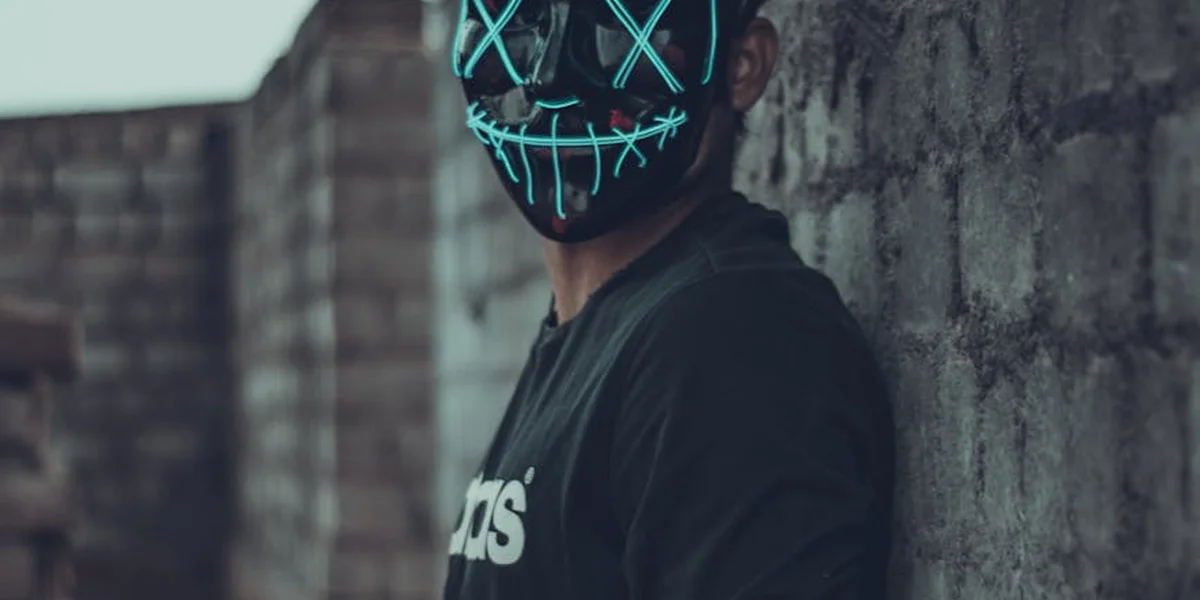 Person wearing a glowing blue stitched mask leans against a brick wall, symbolizing change and the tension between concealment and revelation.