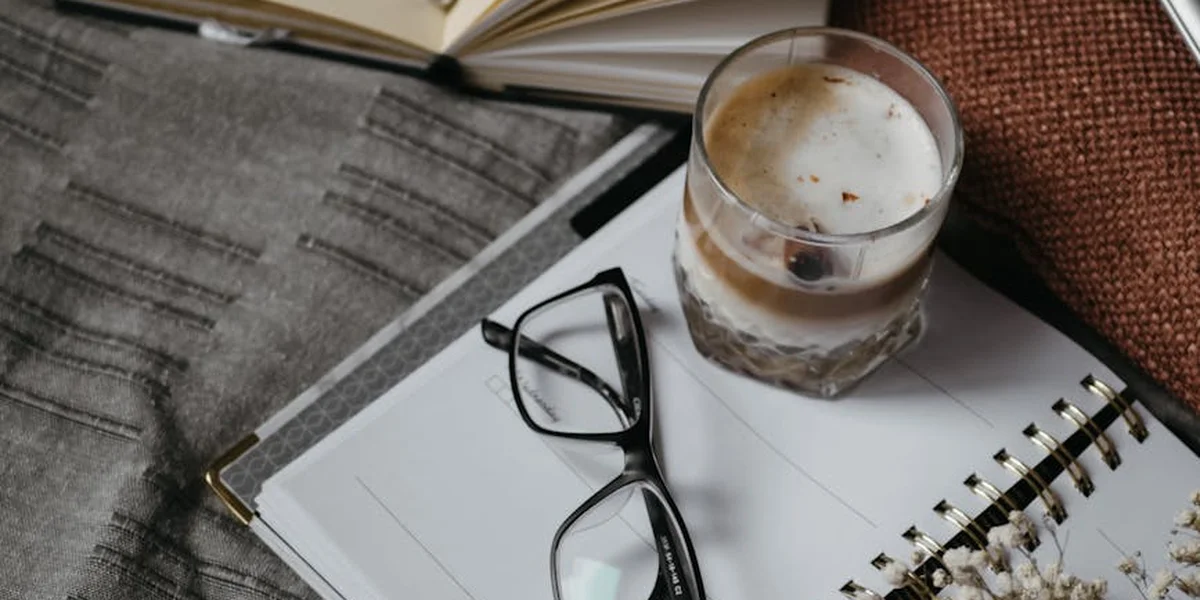 A desk scene featuring a spiral notebook with black-rimmed glasses resting on top, an open book in the background, and a glass with frothy drink nearby.