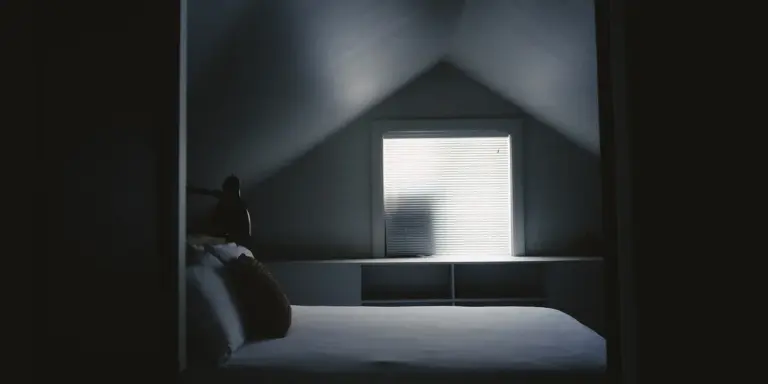 Dim attic bedroom with a single bed and a window glowing softly, the slanted ceiling and shadows conveying isolation and unease.