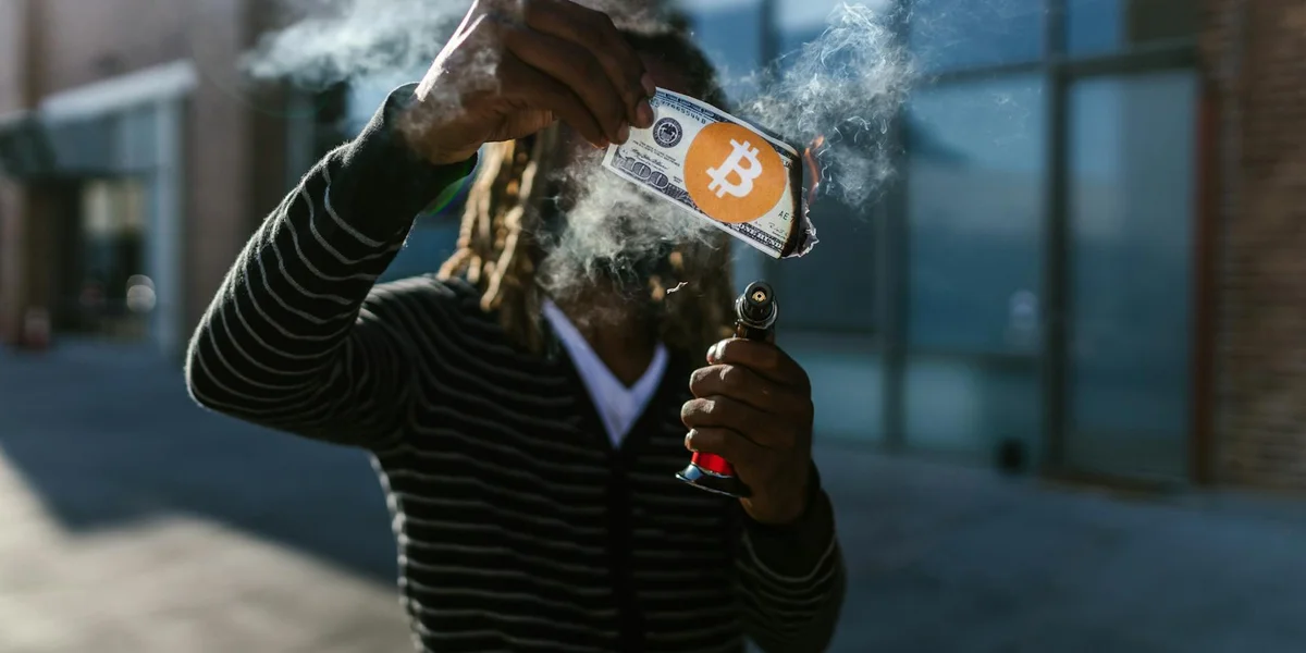A person wearing a striped hoodie stands in a dim urban night setting, holding a lighter with smoke and a card displaying a Bitcoin symbol, evoking robbery imagery.