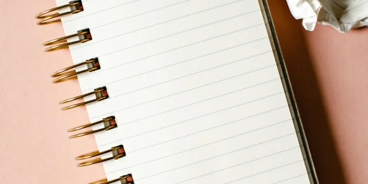 A spiral-bound notebook with blank lined pages lies on a peach-colored surface, with a crumpled piece of paper in the corner.
