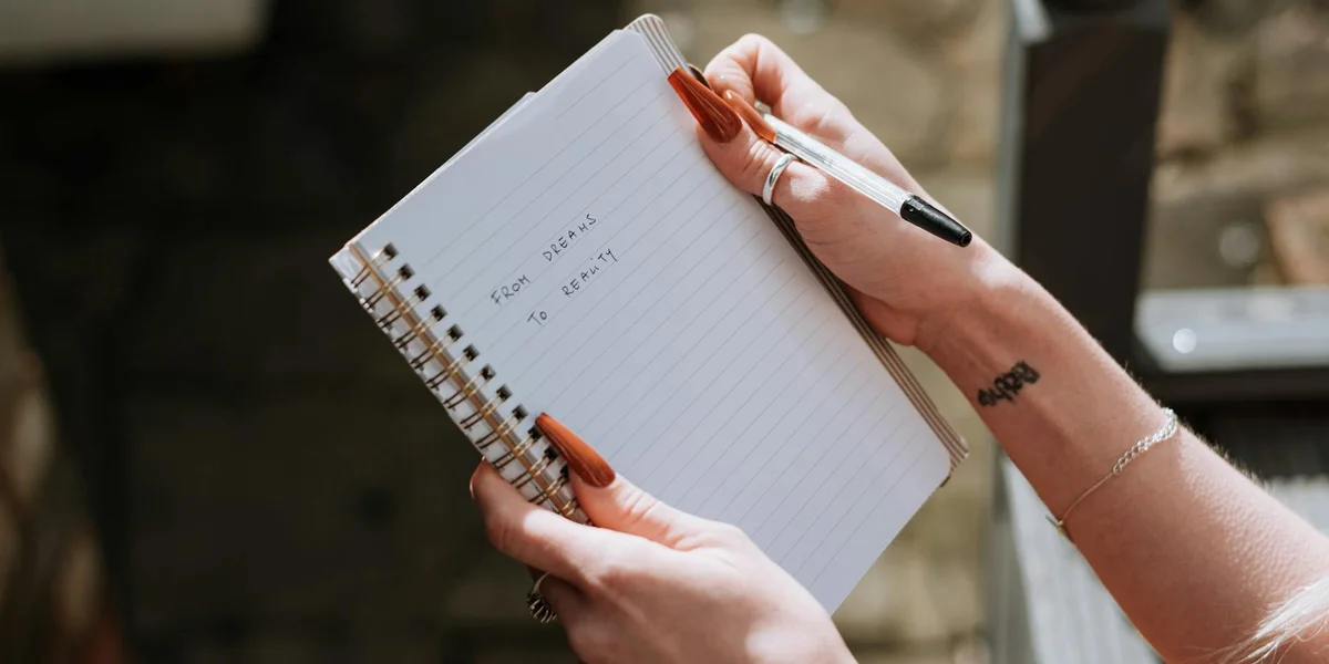 Close-up of hands writing in a spiral notebook with a pen, wearing bracelets and rings.