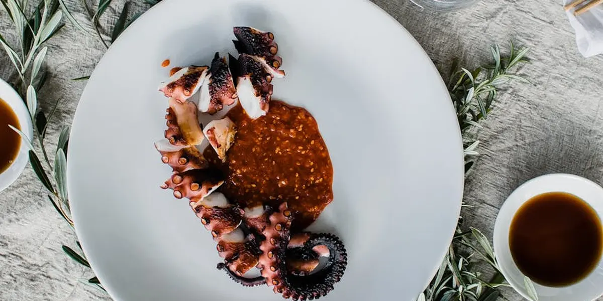 A white plate holds cut octopus tentacles with a dollop of reddish-brown sauce, set on a textured surface with herbs and small cups nearby.