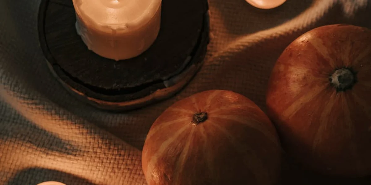 Two orange pumpkins and a lit candle on a textured fabric, bathed in warm orange light.