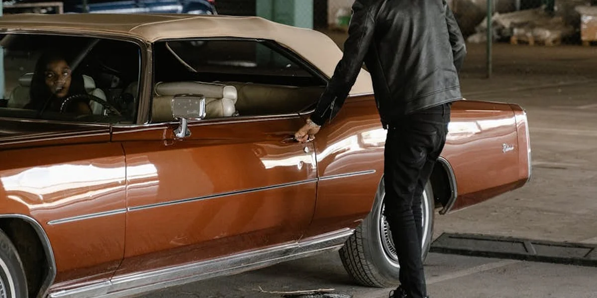 A person in a dark jacket stands beside an orange classic car in a dimly lit parking garage.