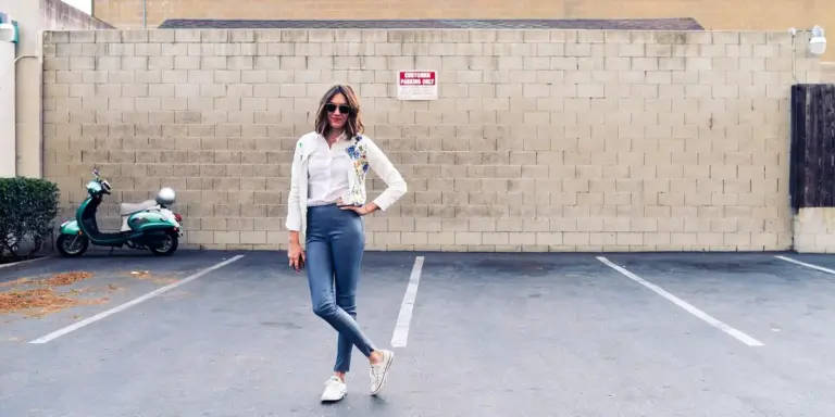 A woman wearing sunglasses, a white jacket, and jeans stands in an empty parking lot in front of a brick wall.