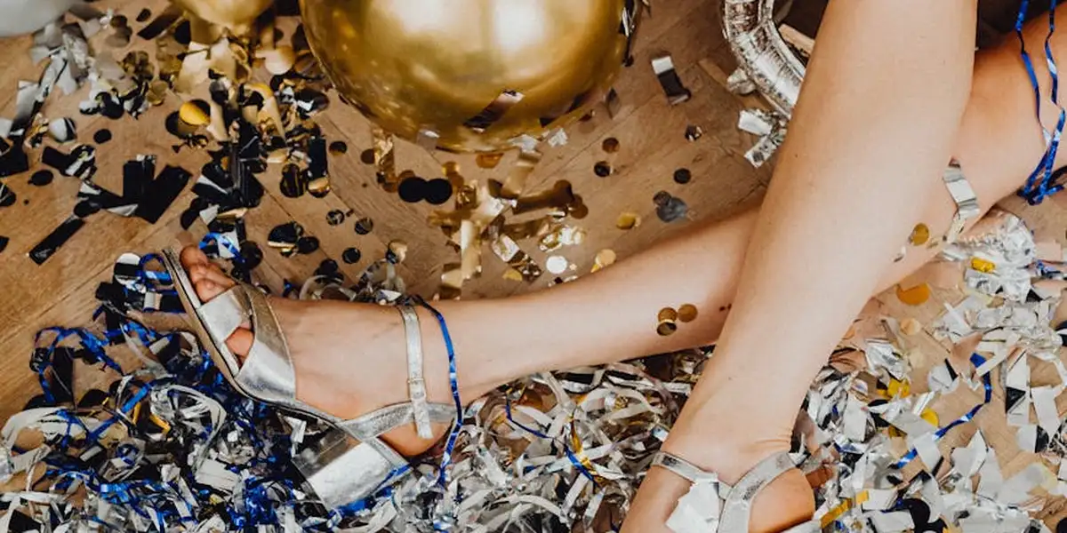 Close-up of legs in silver sandals among scattered party confetti and a gold balloon, suggesting a festive scene with an uneasy mood.