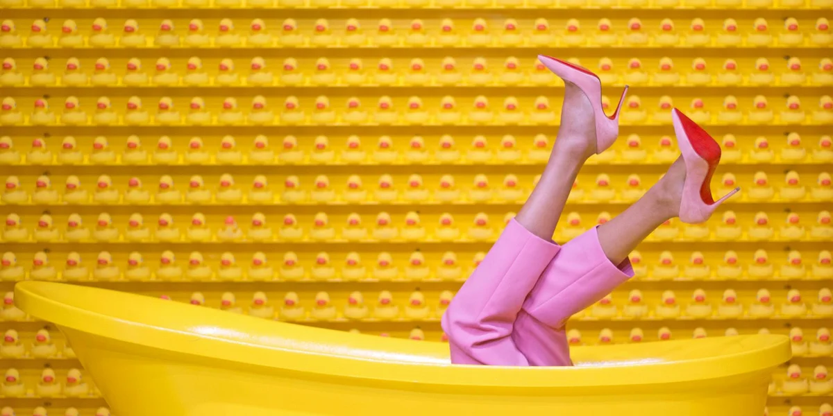 A person wearing pink pants and pink high-heeled shoes with legs raised in a bright yellow tub against a vivid yellow backdrop.