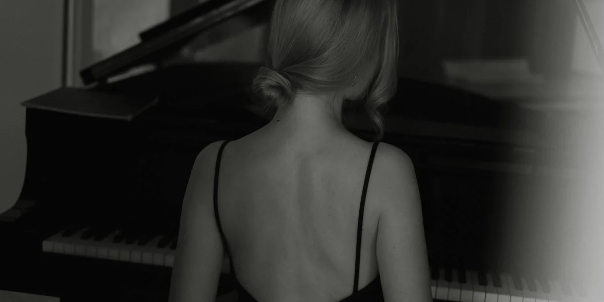 Back view of a person in a sleeveless top seated at a grand piano, captured in black and white.
