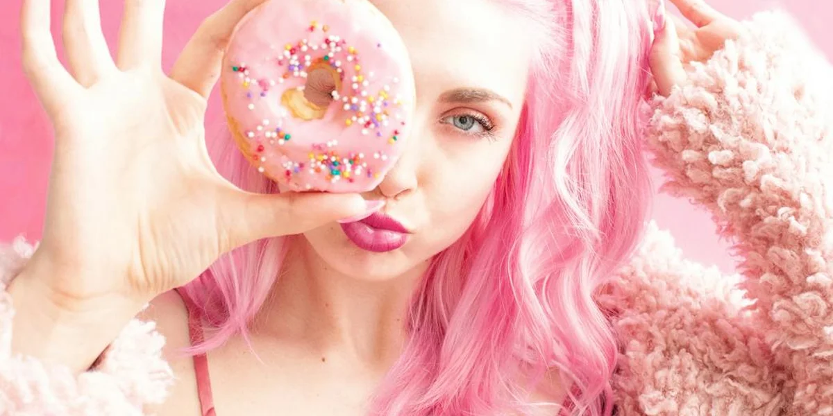 Person with pink hair holding a pink frosted donut up to the camera
