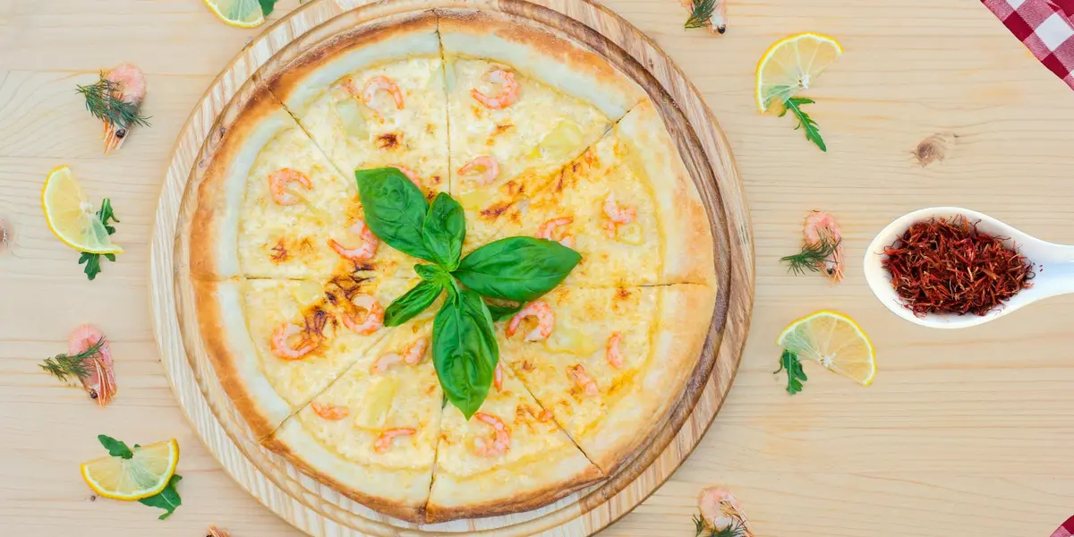 Cheese pizza on a wooden serving board with fresh basil, surrounded by lemon wedges and a small bowl of red spice.
