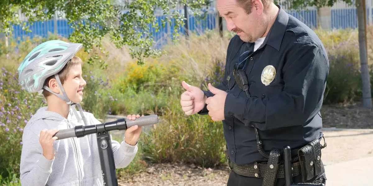 A police officer speaks with a child wearing a bicycle helmet outdoors, in a park setting.