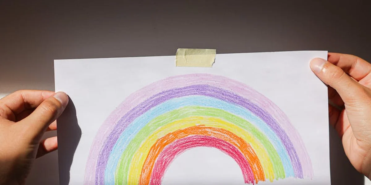 A hand-drawn rainbow on white paper held by two hands, with tape at the top, featuring red, orange, yellow, green, blue, and purple colors.