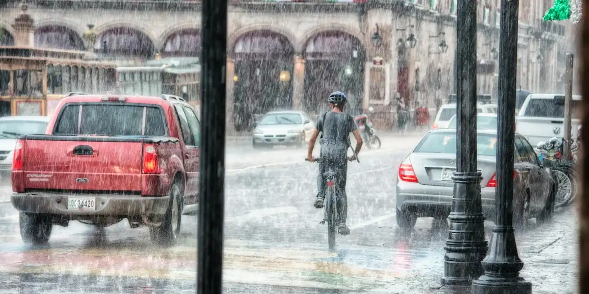 A person riding a bicycle through a heavy rain on a busy city street, with cars and storefronts in the background