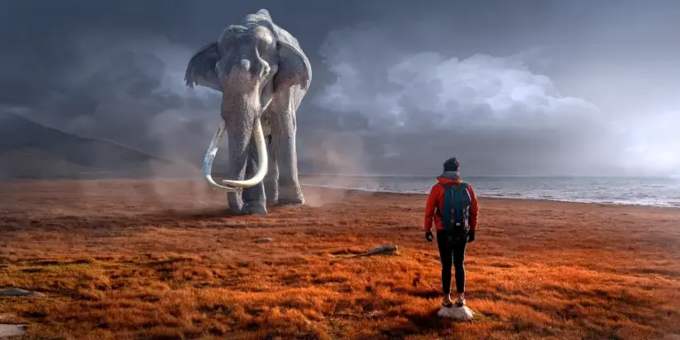 A person in a red jacket with a backpack stands on a desolate, reddish landscape by the water, facing a colossal elephant-like creature with long tusks under a cloudy sky.