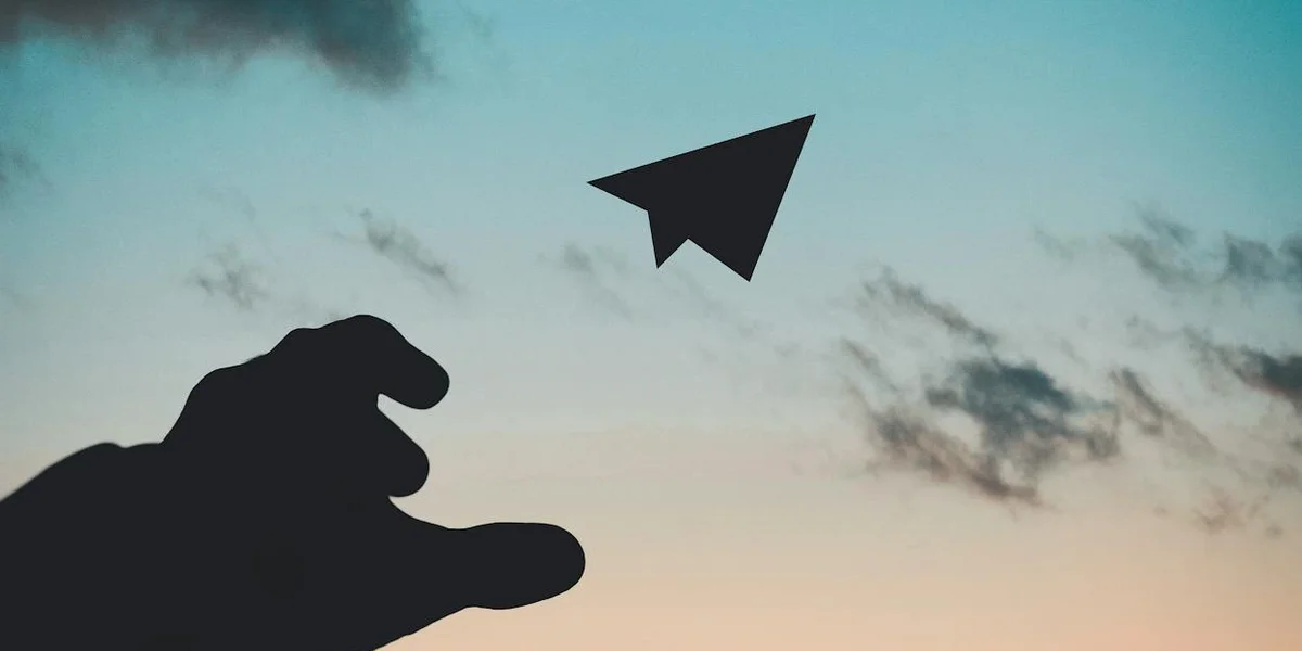 A silhouetted hand reaching toward a small paper airplane in a blue-to-pale sunset sky.