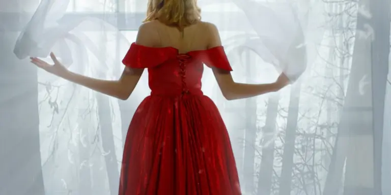 Back view of a person in a red off-the-shoulder dress, standing with arms extended beside sheer white curtains, with light and trees visible outside.