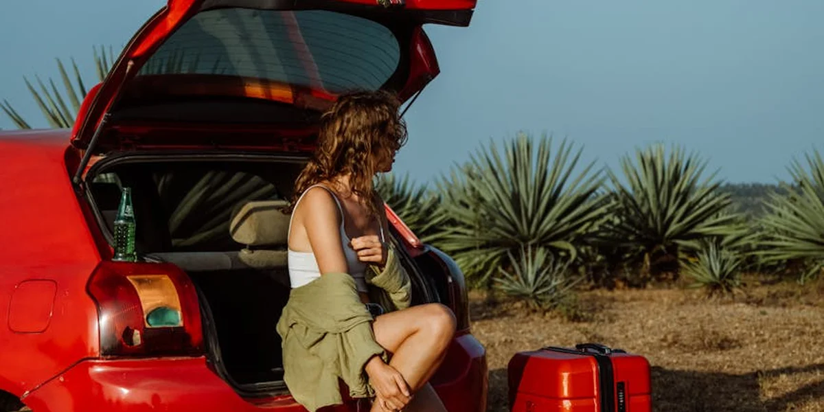A person with curly hair sits in the open trunk of a red car in a desert landscape with spiky desert plants in the background.