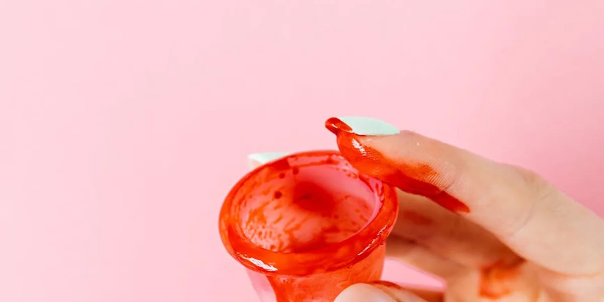 Close-up of a hand holding a small red container with menstrual blood against a soft pink background.
