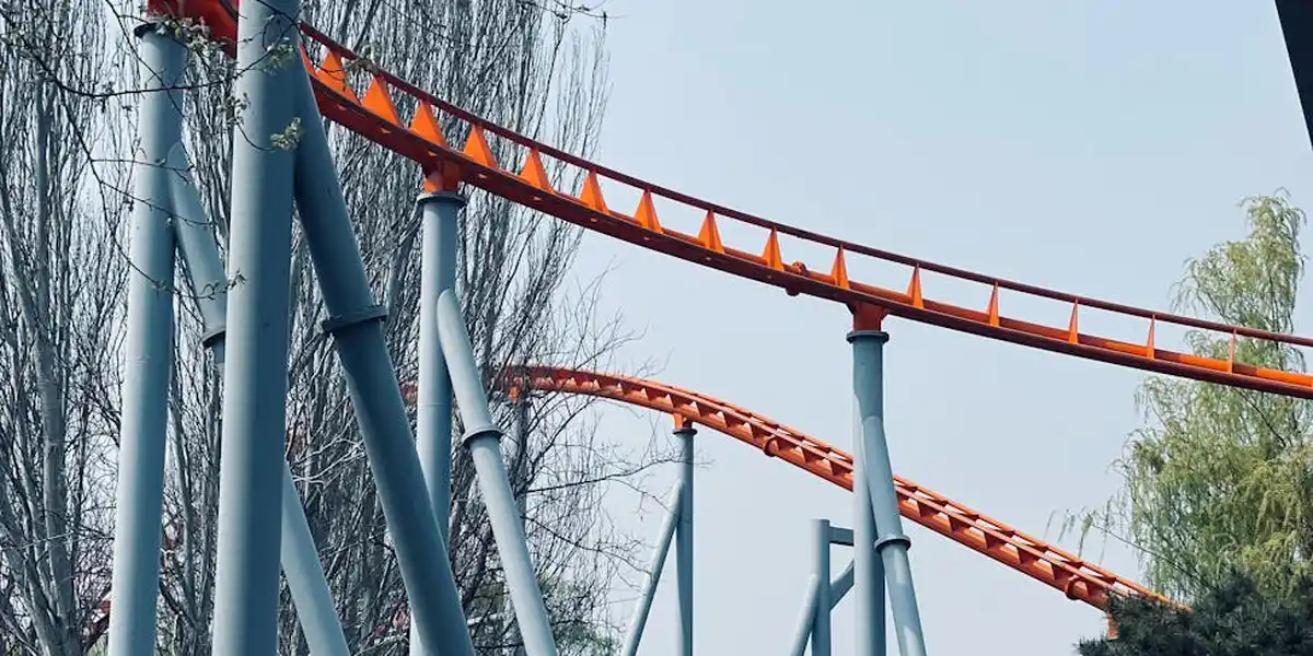 Blue roller coaster supports and orange track twisting against a pale blue sky with bare trees in the background.