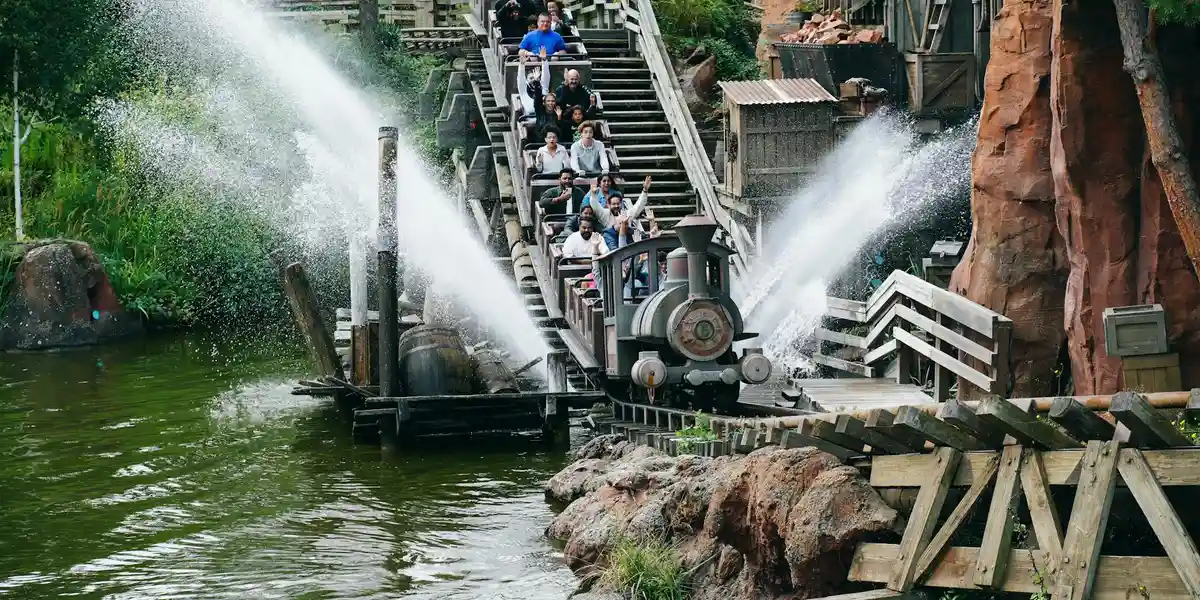People on a wooden log-flume ride as water sprays from jets, descending toward the water with rocky scenery around.