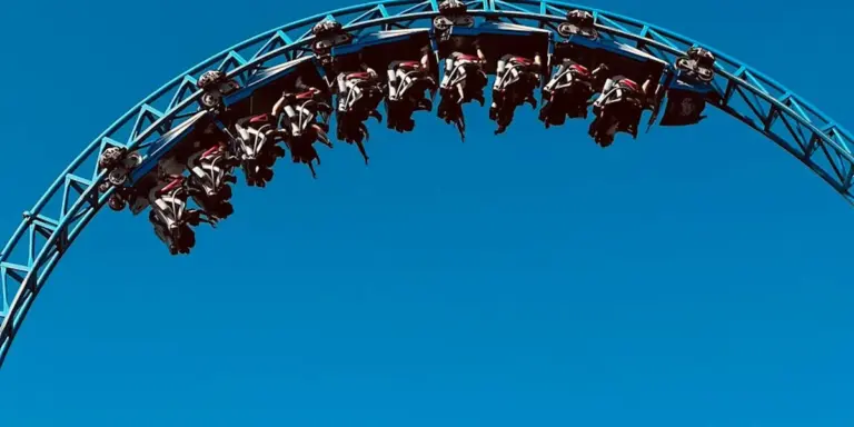A steel roller coaster car train loops along a curved track against a clear blue sky, with riders visible in the cars.