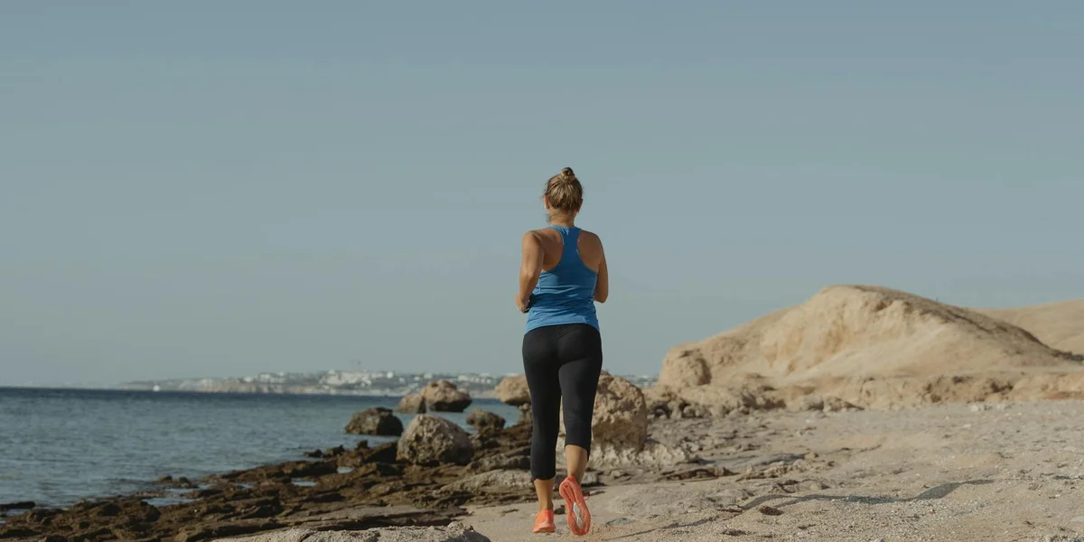 A person in blue athletic wear runs along a rocky beach with sandy dunes on the right and a calm sea on the left, under a clear blue sky.
